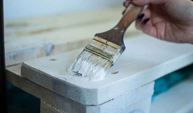 A hand painting a piece of wooden outdoor furniture with white paint.