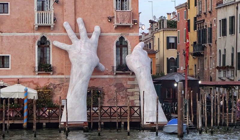 Large models of human hands emerging from a Venice canal holding an orange building.